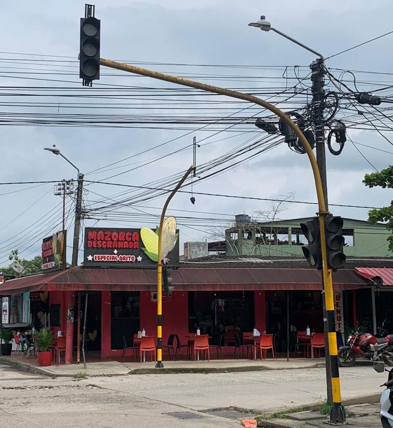 Cruce vehicular en Orito con postes amarillos y cabezas de semáforo apagadas, cables aéreos y locales comerciales alrededor.