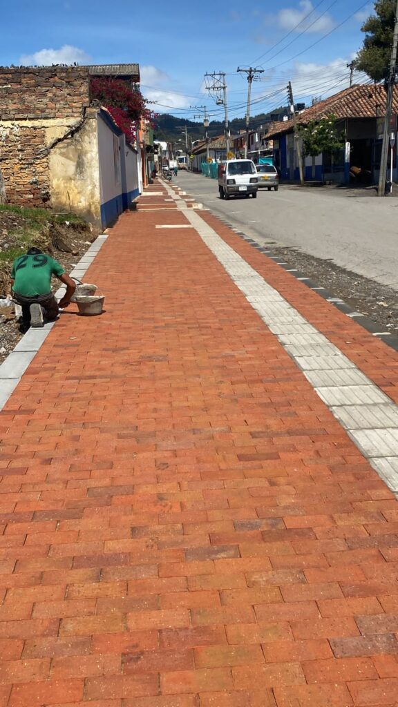 Vista de la Calle 9 de Simijaca, con andén de adoquín rojo y franja podo‑táctil, vehículos circulando y viviendas tradicionales al fondo.