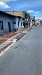 Vista lateral del andén completo en la Calle 9 de Simijaca, adoquines rojos uniformes con franjas podo‑táctiles grises, bordillo de concreto y fachada de color azul y verde de viviendas adyacentes, bajo un cielo despejado.