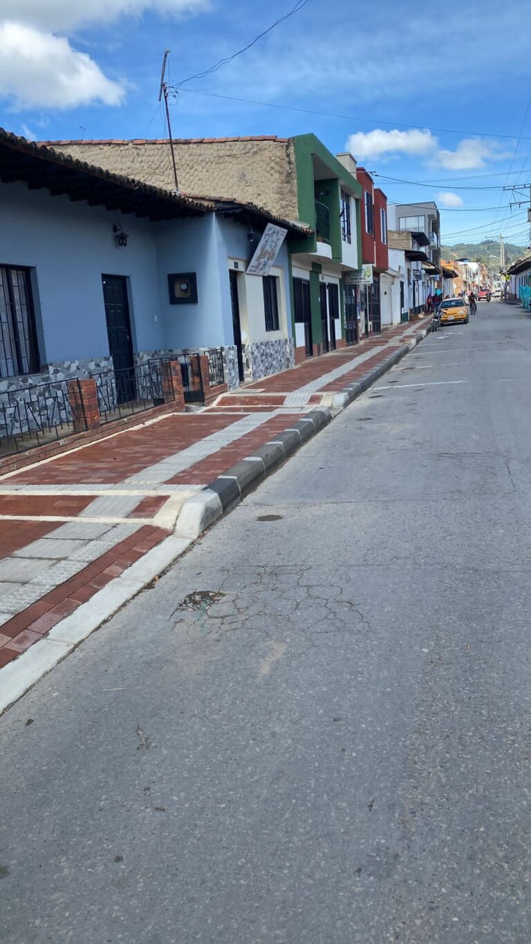 Vista lateral del andén completo en la Calle 9 de Simijaca, adoquines rojos uniformes con franjas podo‑táctiles grises, bordillo de concreto y fachada de color azul y verde de viviendas adyacentes, bajo un cielo despejado.