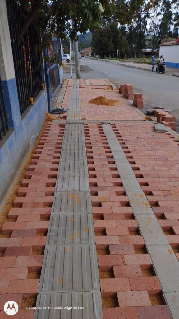 Andén nuevo revestido con adoquines rojos y franja central de baldosas podo-táctiles grises, frente a fachada blanca con arco y un perro caminando.