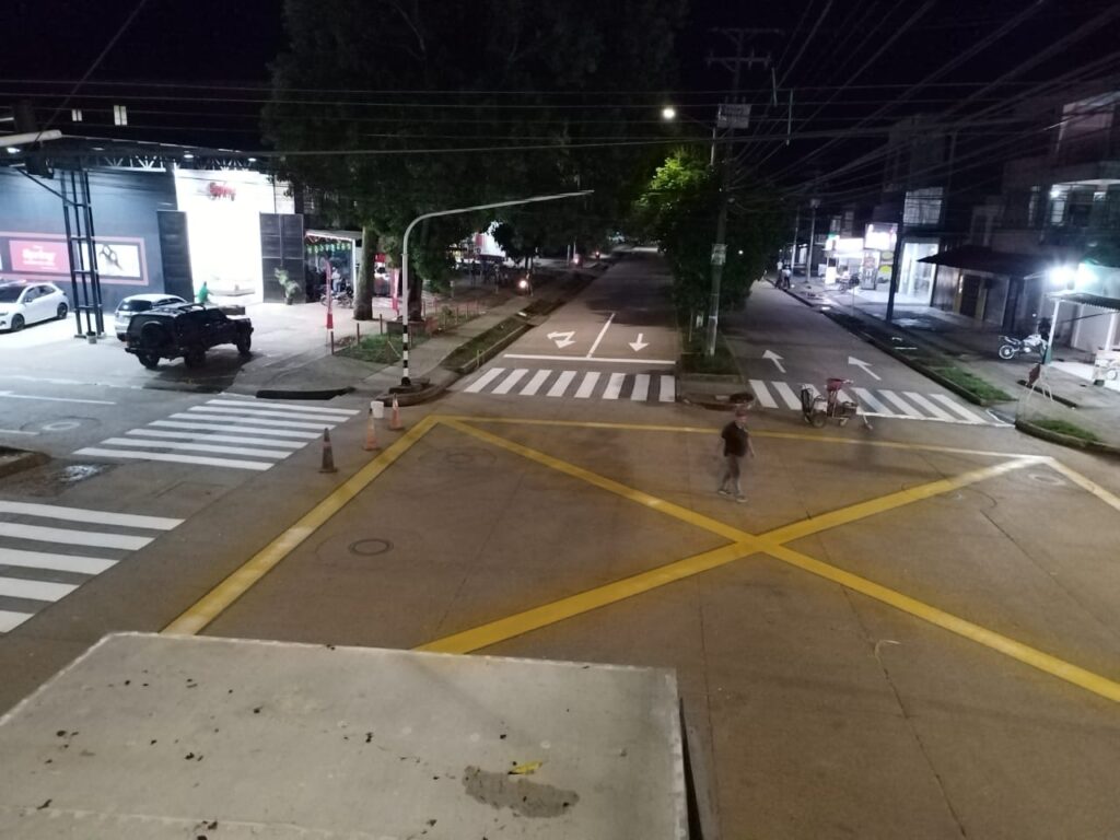 Vista nocturna de una intersección en Sesquilé con línea amarilla de “caja paraguas” en el pavimento, cebras de paso peatonal en cada brazo, flechas direccionales y bordillos elevados. Al fondo, locales comerciales iluminados y un gabinete de control semafórico abierto con un computador portátil conectado, reflejando tareas de revisión y ajuste de la señalización vial.