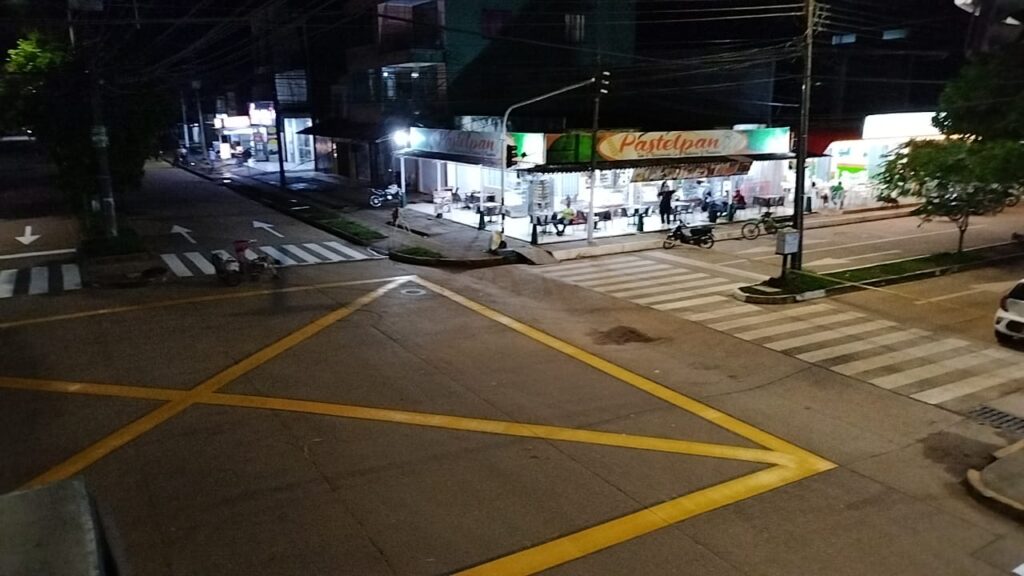 Intersección de noche con demarcación en “caja paraguas” amarilla, cebras peatonales blancas, flechas direccionales y aceras elevadas; al fondo, locales comerciales iluminados.