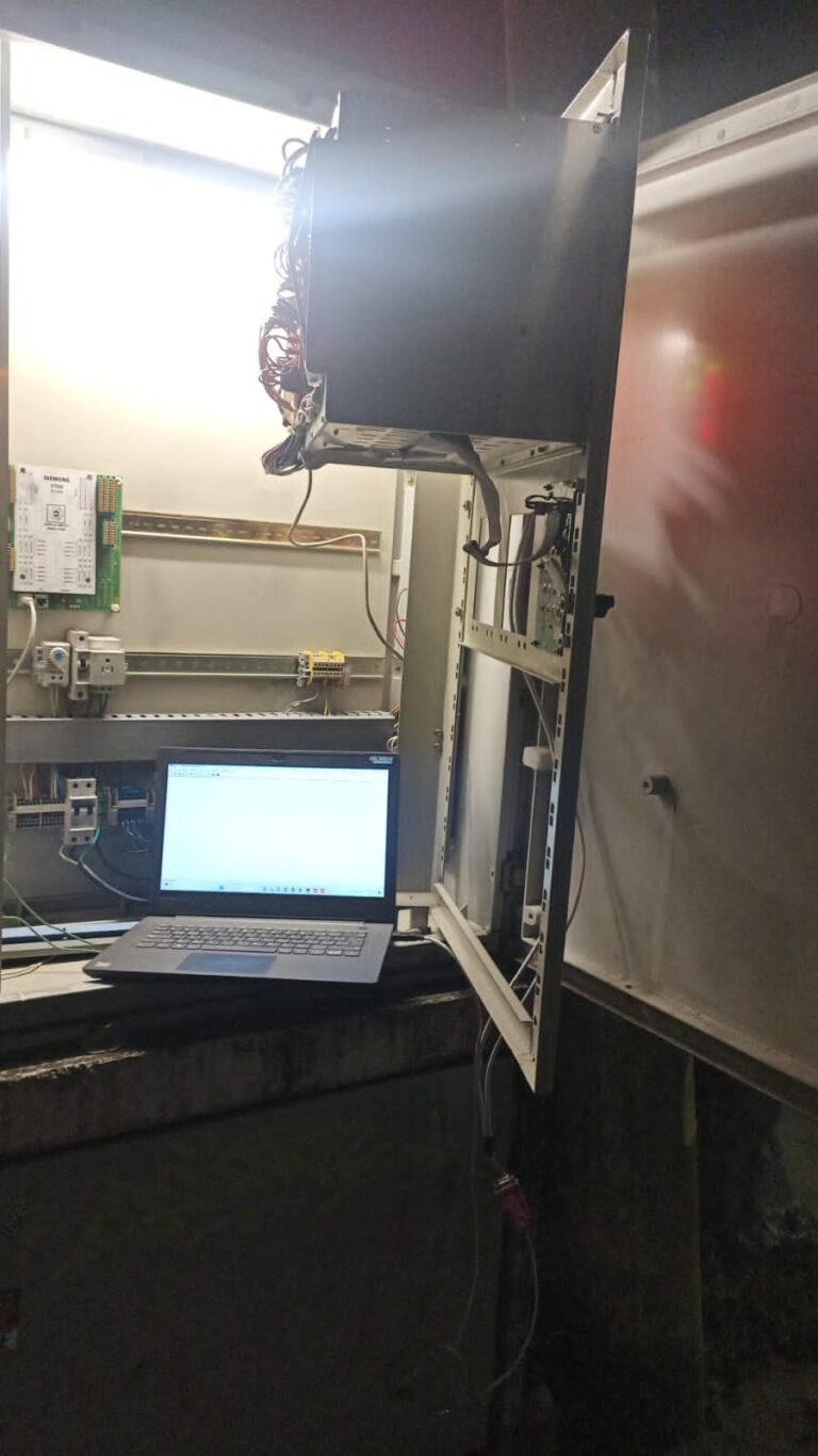 Interior de un gabinete semafórico abierto de noche, con módulos electrónicos Siemens montados en riel DIN. Un portátil está conectado dentro del gabinete, mostrando software de parametrización de tráfico. Cables y terminales se ven organizados, y luces de estado verdes y rojas indican alimentación y señalización activas.