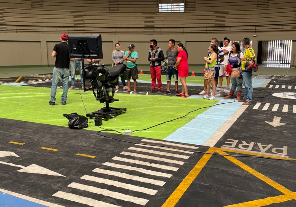 Grupo de ciudadanos frente a pantalla y simulador de motocicleta sobre pista con demarcación vial dentro de coliseo en Granada Meta.