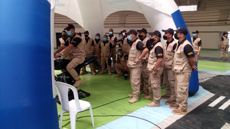 Grupo de uniformados observa a participante en simulador de motocicleta dentro de carpa educativa de seguridad vial.