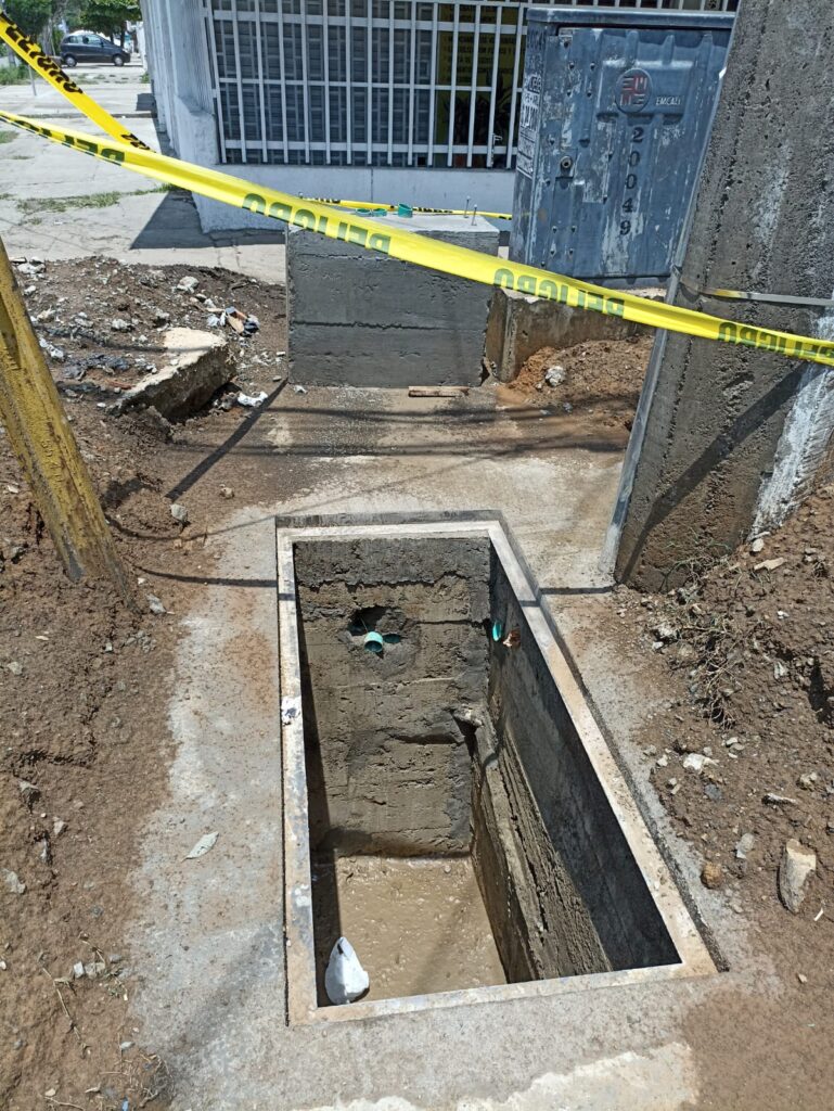 Zanja de concreto abierta con tuberías y caja de inspección señalizada con cinta de peligro en vía urbana de Cali.