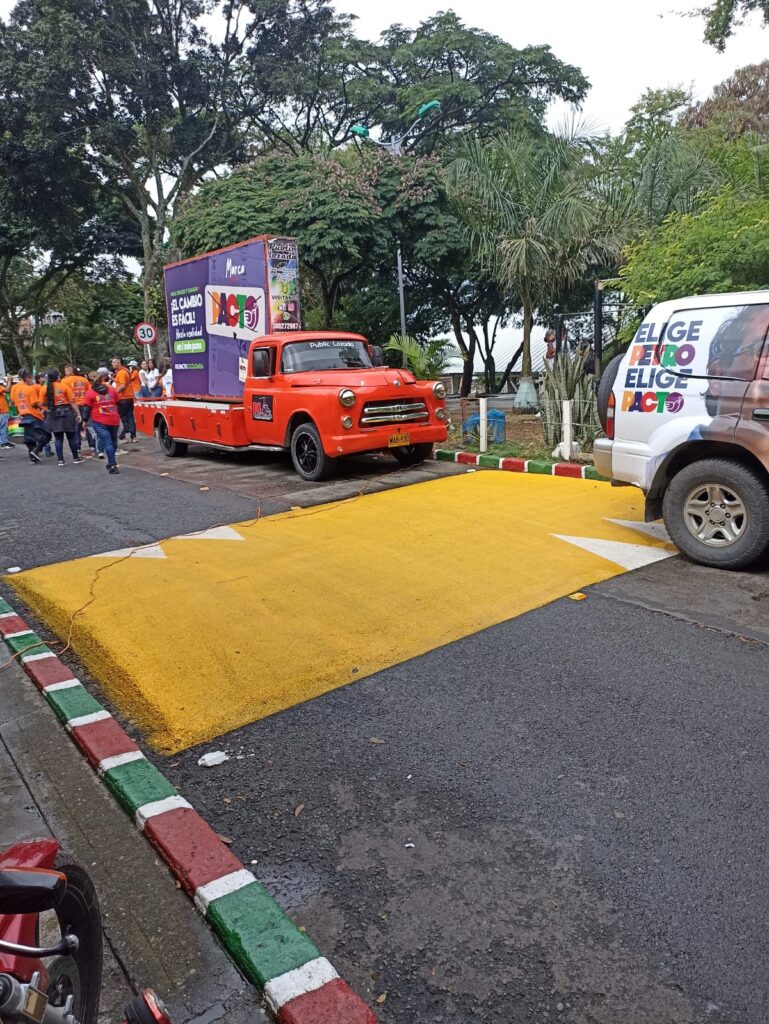 Loma reductora recién pintada en amarillo con flechas blancas, camioneta y camión publicitario; equipo de interventoría vial en vía urbana de Cali.