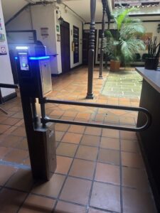 Torniquete de un solo carril con escáner facial y barrera de acero inoxidable, junto a un patio interior ajardinado.
