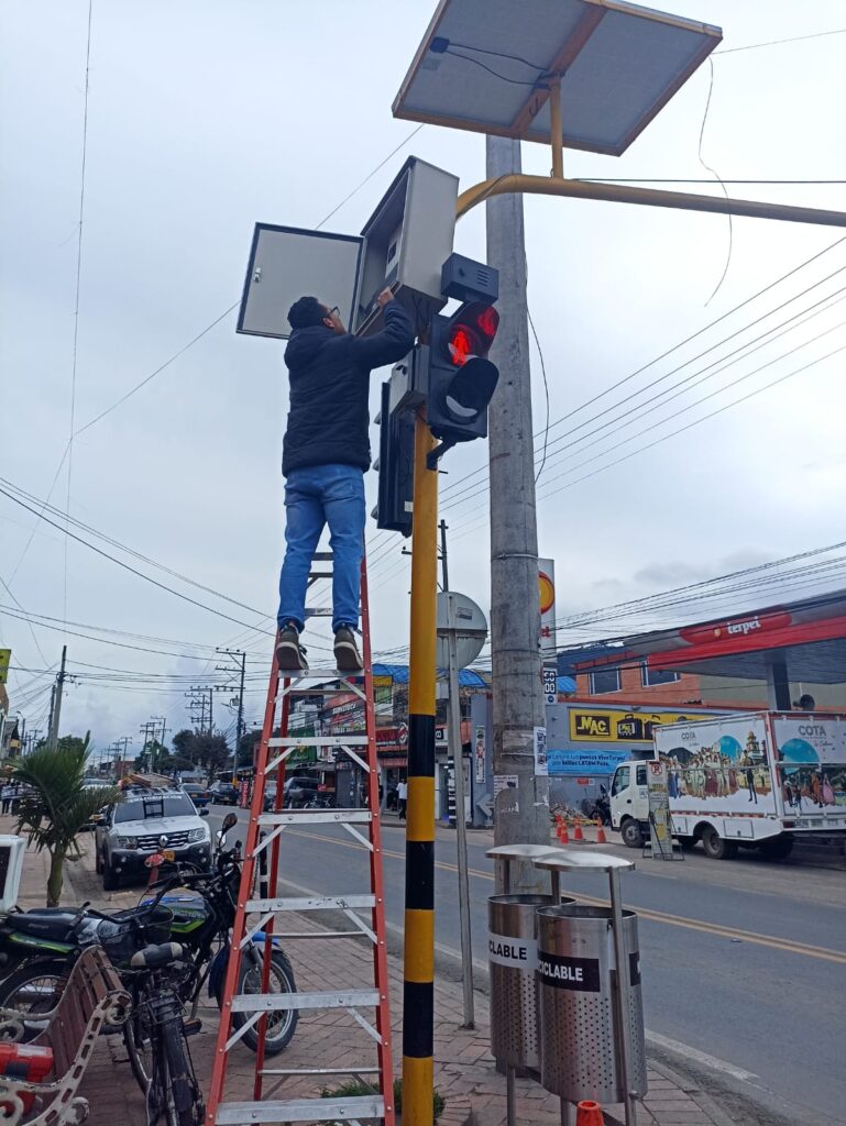Técnico en escalera instala semáforo con panel solar y gabinete de control en intersección urbana.