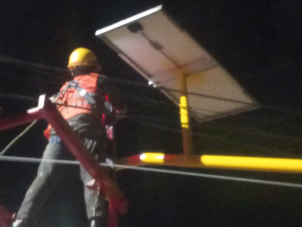 Técnico con casco y chaleco reflectante en andamio realizando mantenimiento nocturno de panel solar y controlador de tráfico inalámbrico.